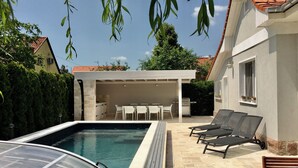 Una piscina con un porche cubierto, sillas de descanso y un coche aparcado cerca.