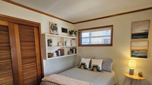 Ein Schlafzimmer mit einem Bett, einem Nachttisch mit Lampe, einem Fenster mit Jalousien, einem Bücherregal und einer Tür mit Holzjalousien.