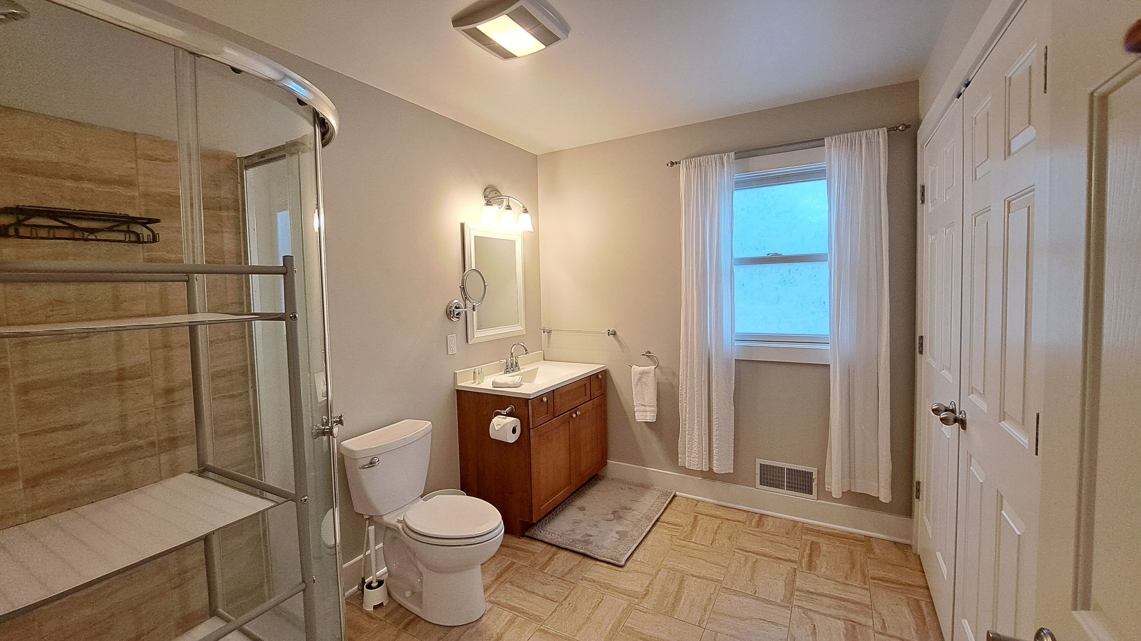A bathroom with a glass shower enclosure, a toilet, a sink with a vanity, and a window with curtains.