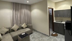 Un salon moderne avec un canapé gris, une table basse et un coin cuisine doté de meubles gris et d’un réfrigérateur noir.