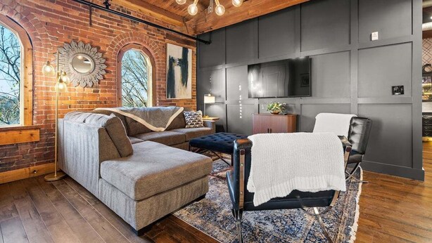 A cozy living room with a sectional sofa, a black leather chair, and a large mirror on the brick wall.
