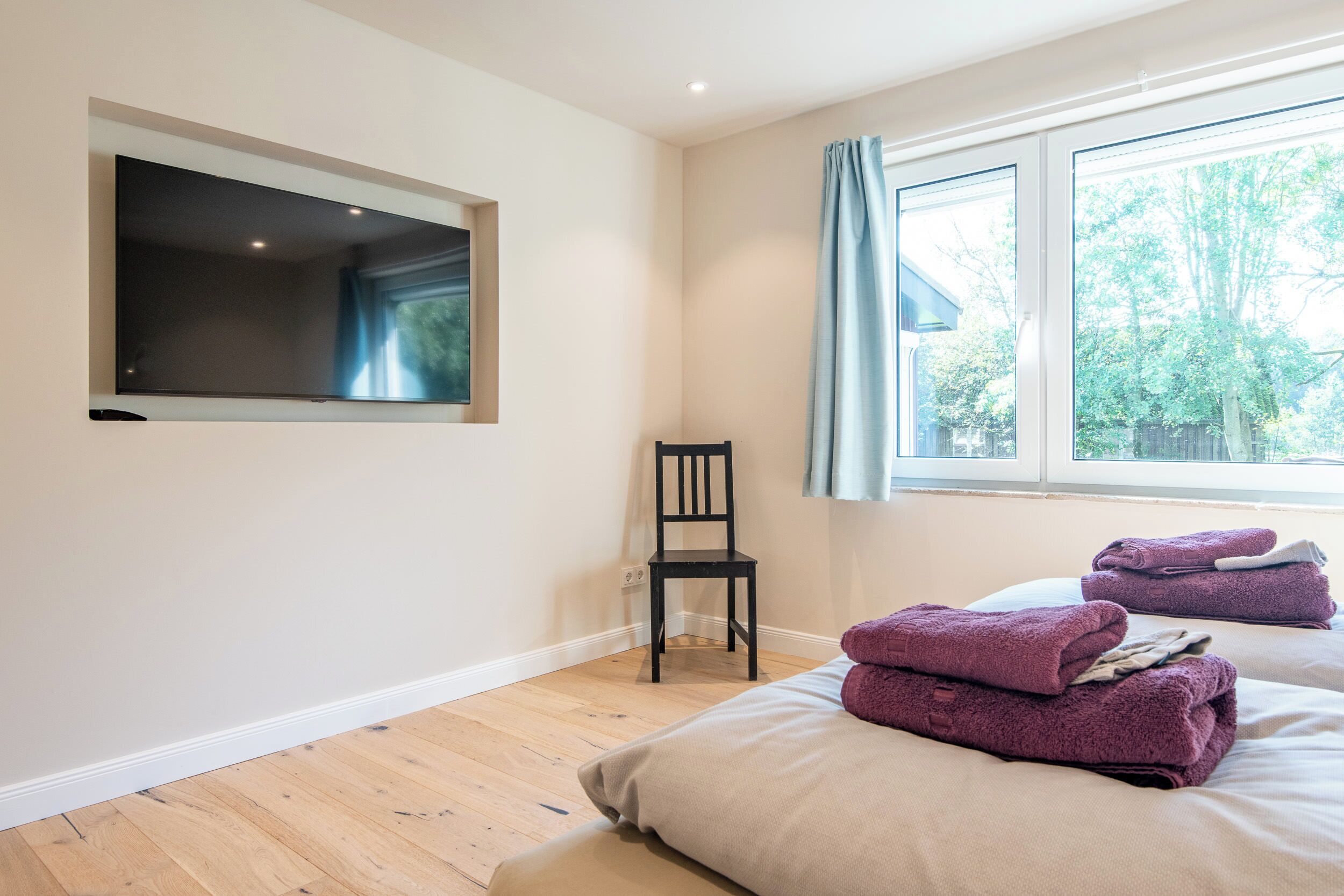 Ein Schlafzimmer mit großem Fenster, einem an der Wand befestigten Flachbildfernseher, einem Holzstuhl und ordentlich gefalteten Handtüchern auf dem Bett.