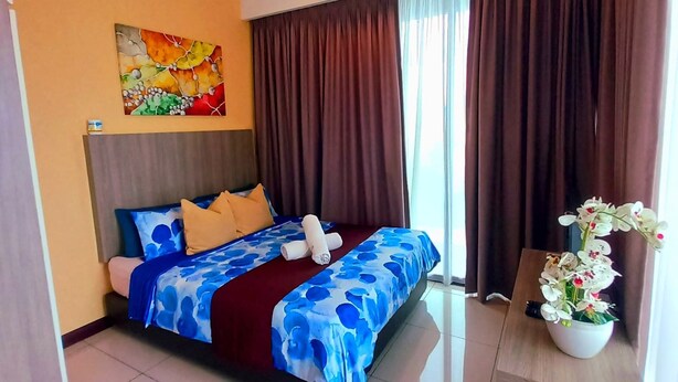 Una habitación de hotel con cama, mesita de noche, ventilador de techo, equipo de aire acondicionado, ventana con cortinas y un cuadro en la pared.