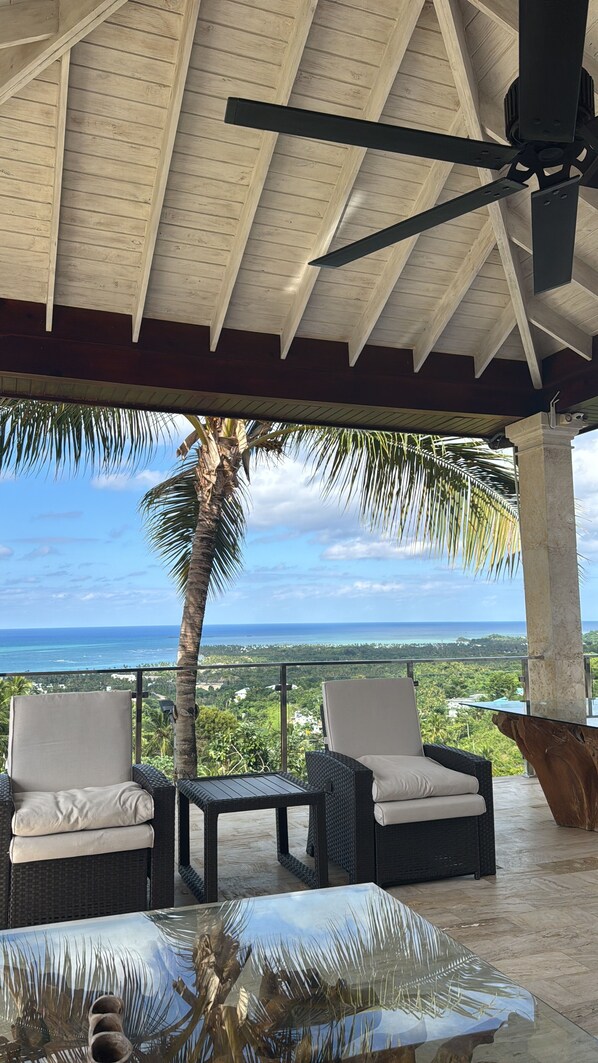 A covered outdoor seating area with two wicker chairs and a glass-top table, overlooking a scenic ocean view.