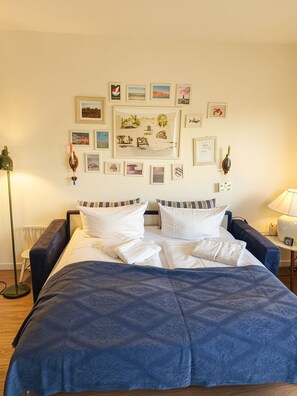 A bedroom with a bed, pillows, a blue blanket, a wall with framed pictures, and a floor lamp.