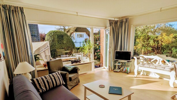 Un salon spacieux avec un canapé, un fauteuil et une table basse. Un téléviseur est fixé sur un meuble, et une plante en pot se trouve dans un coin.