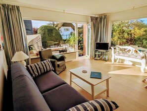 A spacious living room with a sofa, armchair, and coffee table. A television is mounted on a stand, and there is a potted plant in the corner. - Haus Hanne Sylt ♥ Apartment with terrace/garden for 2 persons (Sylt)