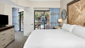 A hotel room with a large bed, a television, a desk with a lamp, and a view of an outdoor pool area.
