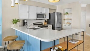 A modern kitchen with white cabinets, a stainless steel refrigerator, a microwave, and a sink. There are two wooden stools and a wine bottle on the counter.