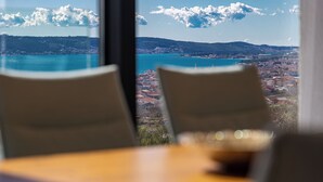 Une vue depuis une fenêtre donnant sur une ville côtière, avec des montagnes à l’horizon et un ciel dégagé.