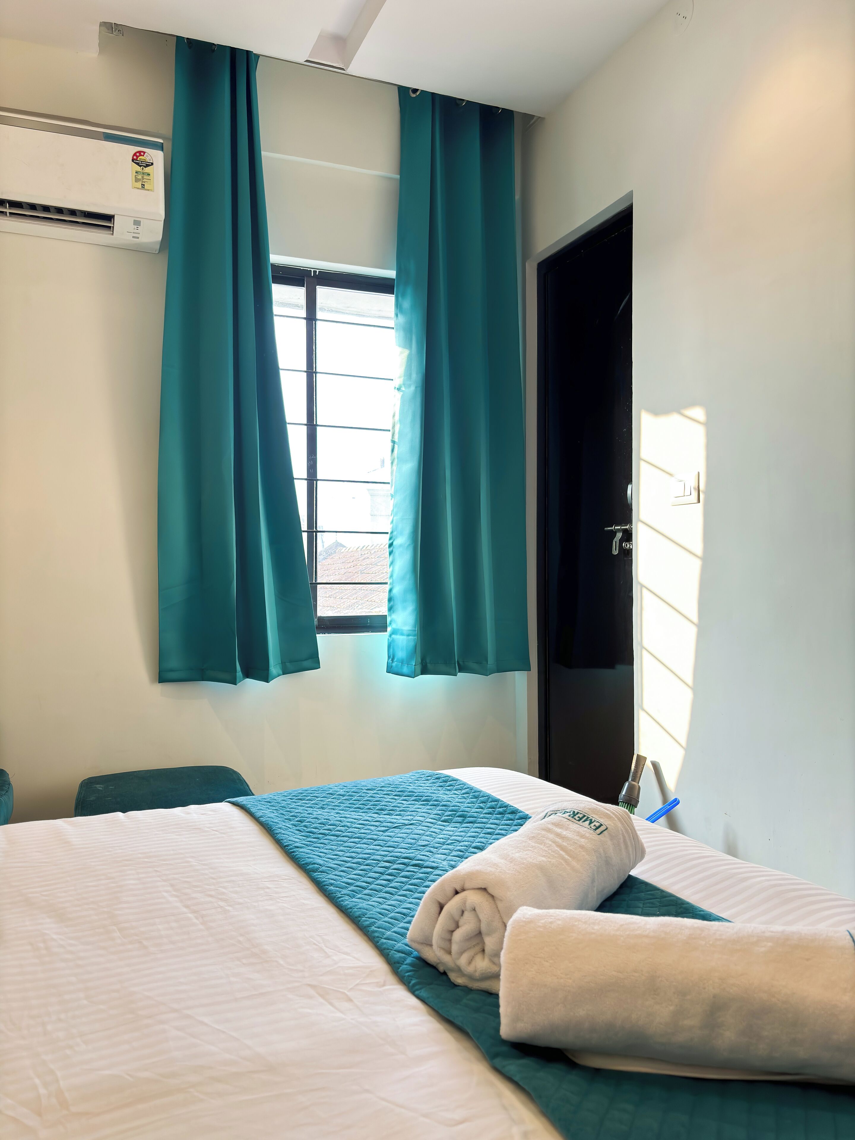A hotel room with a bed, teal curtains, a window, and an air conditioning unit.