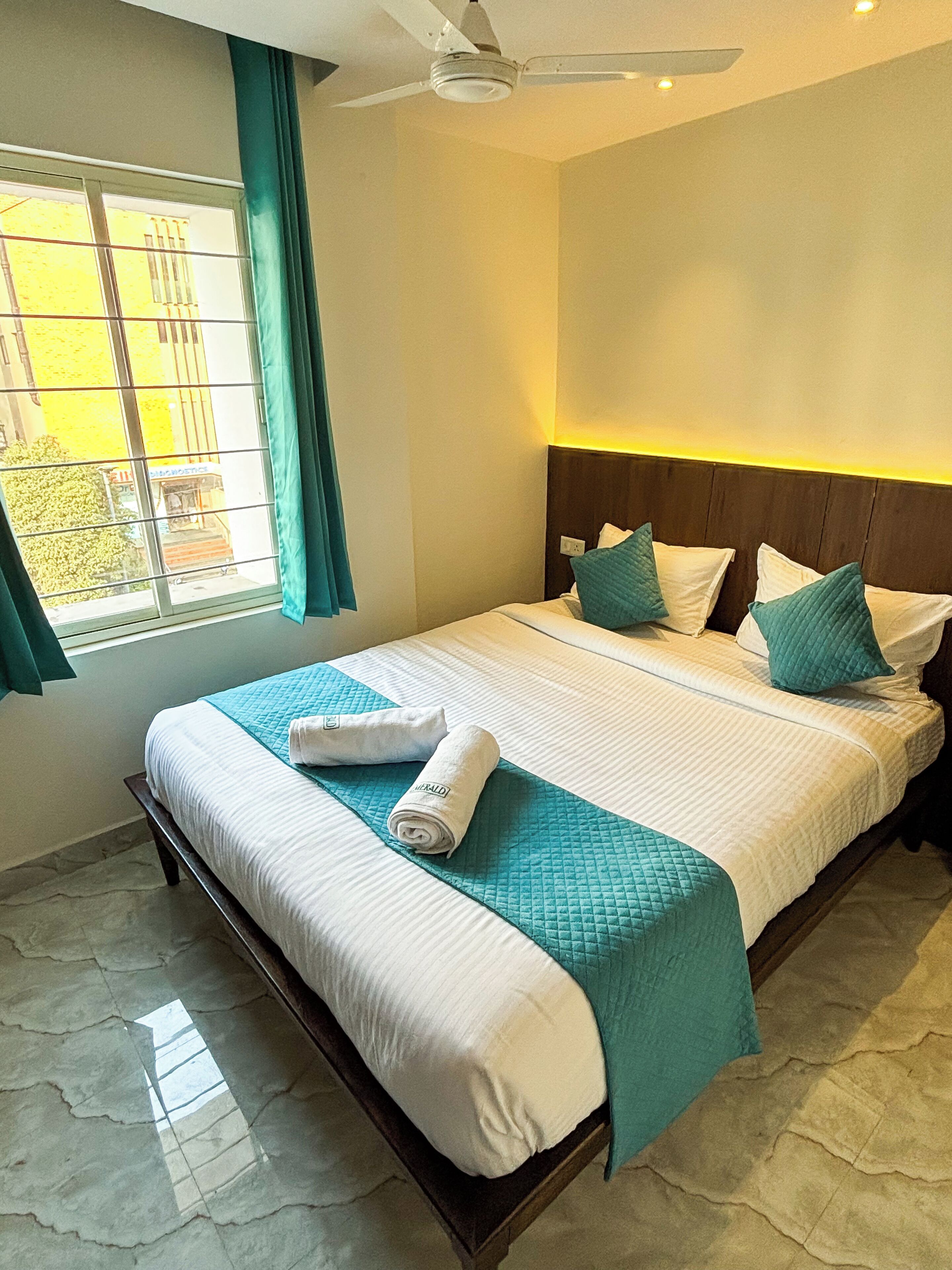 A neatly made bed with a teal quilt, white linens, and a rolled towel on the side.