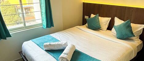 A neatly made bed with a teal quilt, white linens, and a rolled towel on the side.
