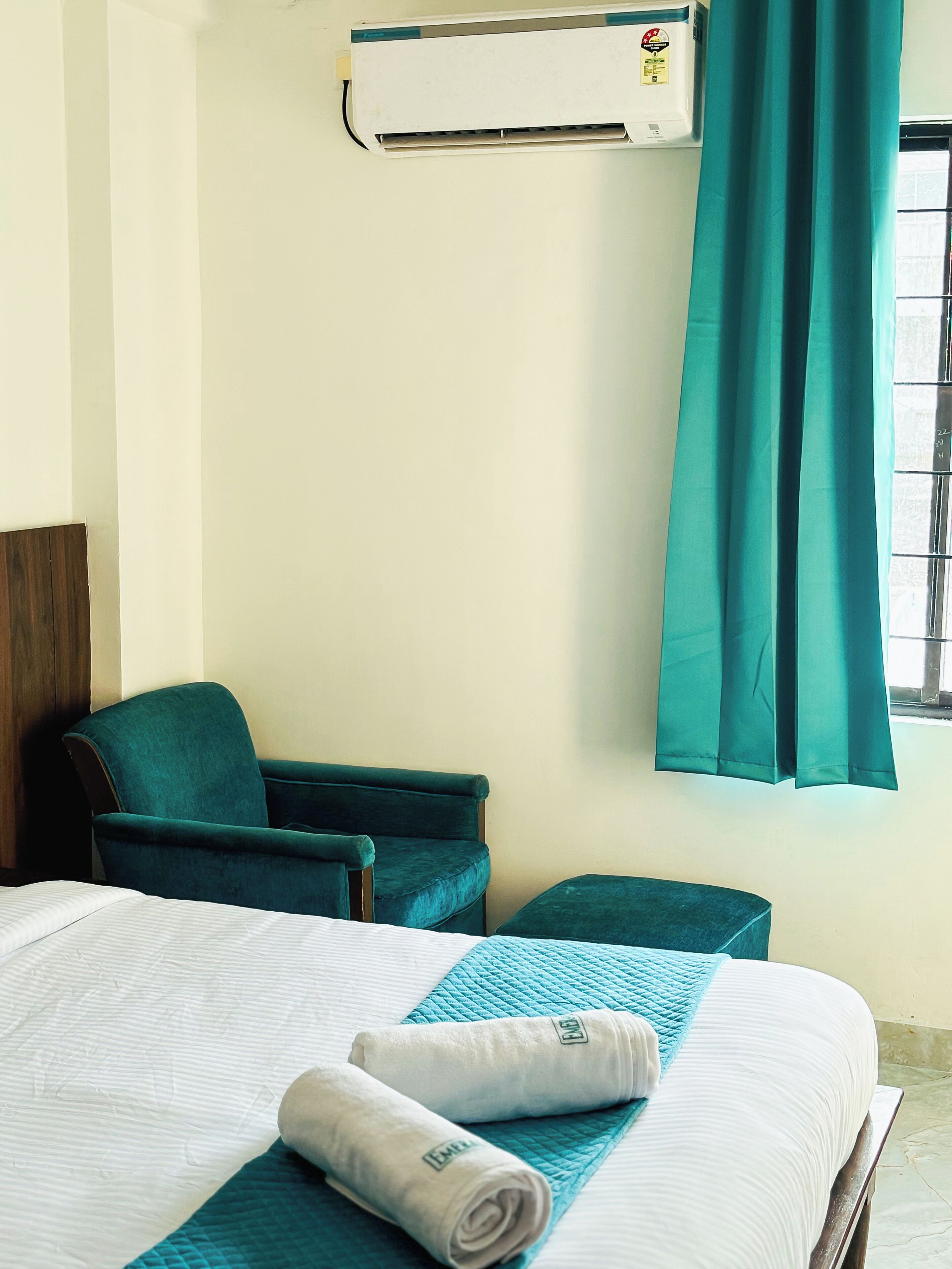 A hotel room with a bed, two chairs, an air conditioning unit, and a window with curtains.