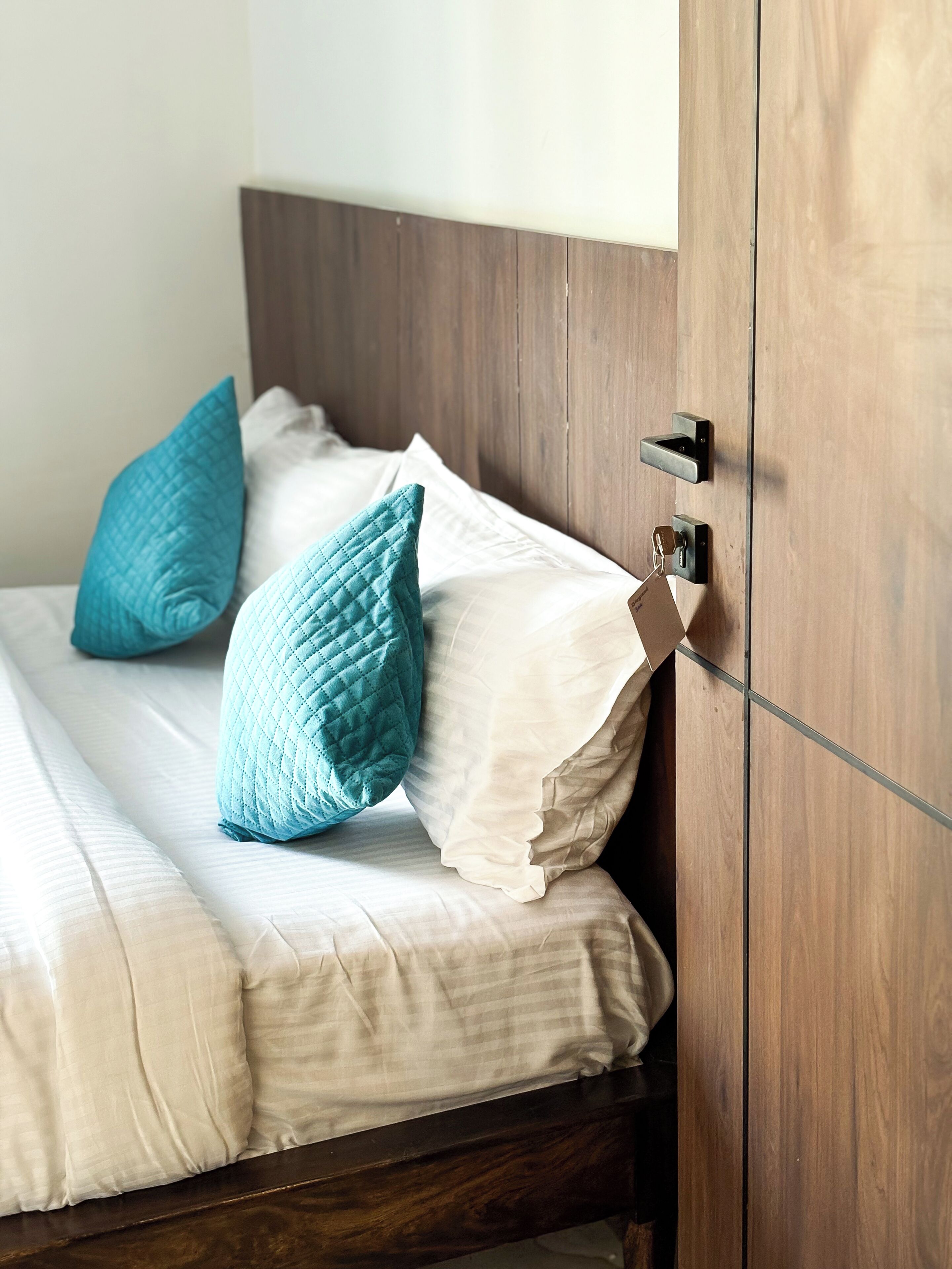 A bed with white linens and teal pillows, a wooden headboard, and a door with a handle and lock.