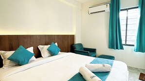 Una camera d'albergo con un letto grande, una testiera in legno, biancheria da letto color verde acqua, un asciugamano arrotolato, una scrivania, una sedia, un condizionatore d'aria e una finestra con tende.