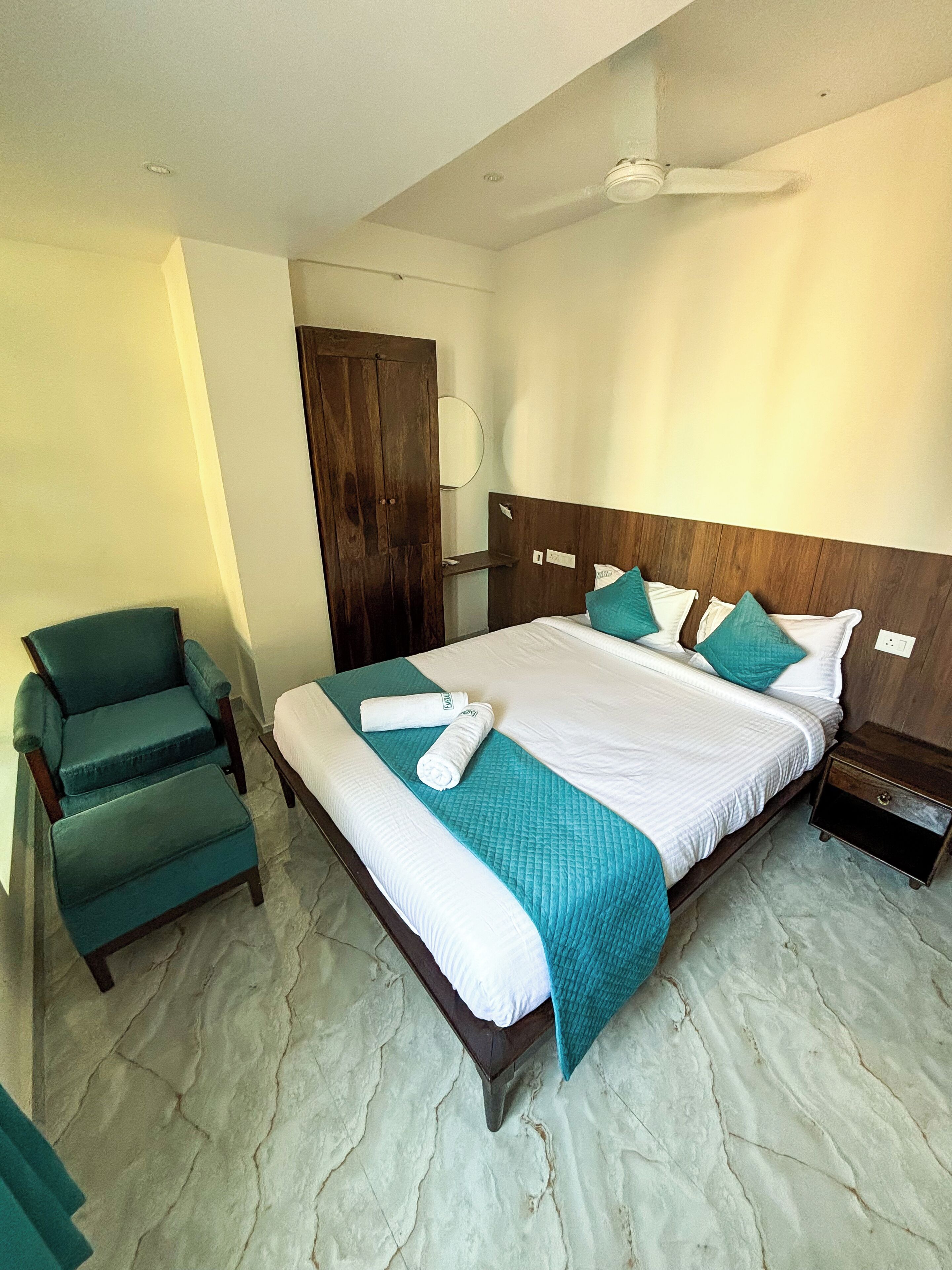 A hotel room with a bed, a chair, a wooden wardrobe, and a ceiling fan.