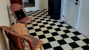 Une pièce avec un sol à carreaux, deux fauteuils en osier, un buffet en bois foncé et une porte blanche avec des fenêtres grillagées.