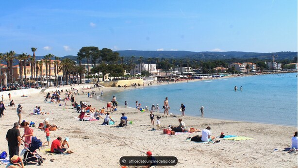 Ein voller Strand mit Menschen, die sich sonnen und schwimmen, Palmen und Gebäuden im Hintergrund.
