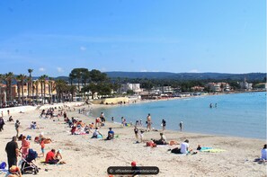 A crowded beach with people sunbathing and swimming, palm trees, and buildings in the background.
