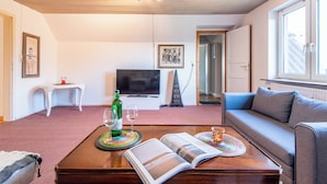 A living room with a sofa, a coffee table with a magazine, a television, and a small table with a bottle and glasses.