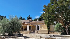 Ein Haus mit Steinwänden und einem Ziegeldach, eine steinerne Treppe führt zum Eingang, und ein Garten mit Olivenbäumen und einem großen Baum.