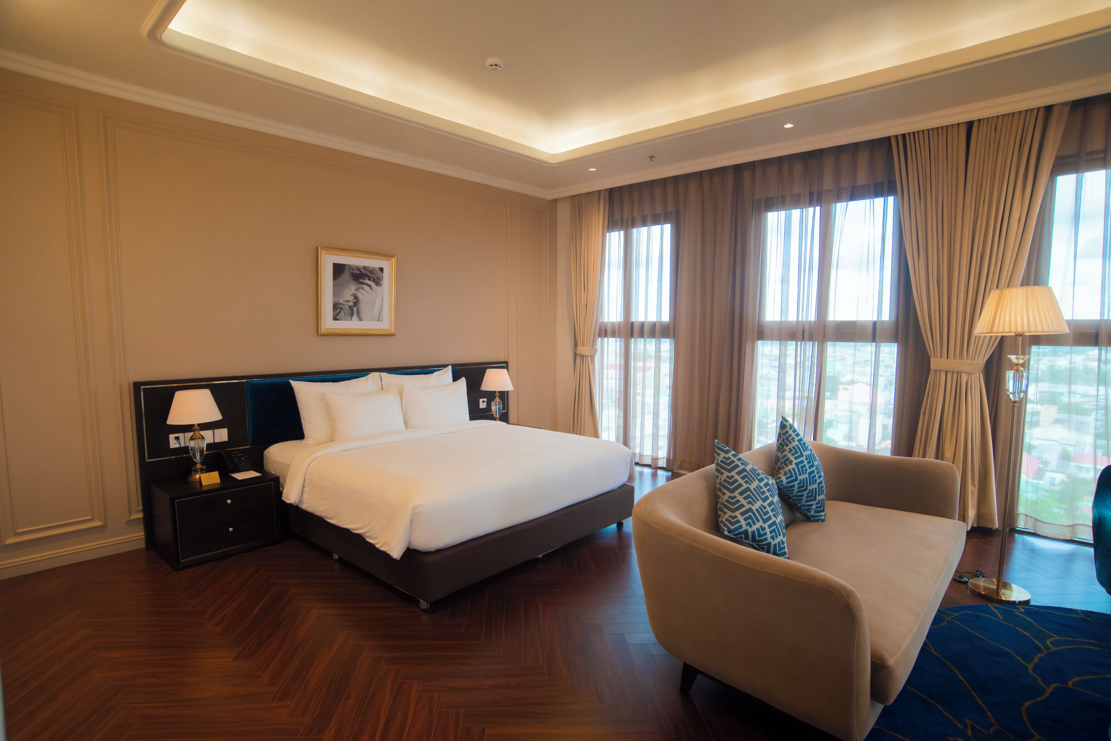 Una habitación de hotel con cama, mesita de noche, lámpara, sofá y ventanas grandes con cortinas.