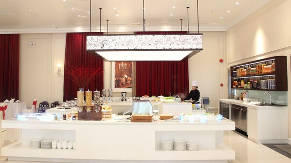 A modern hotel breakfast area with a central counter displaying various food items, a chef preparing food, and a beverage bar on the right.
