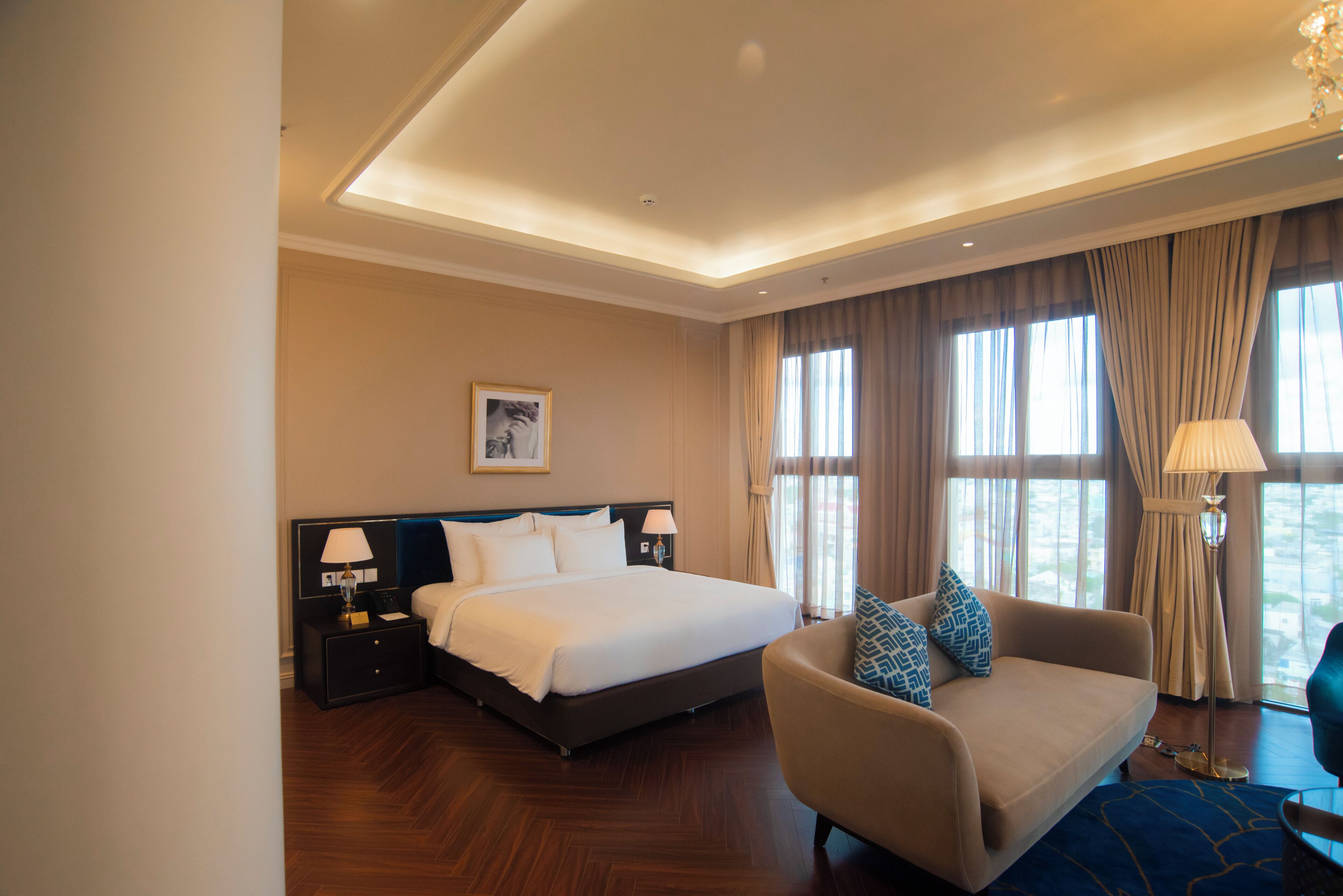 A hotel room with a bed, a sofa, a nightstand with a lamp, and a large window with curtains.