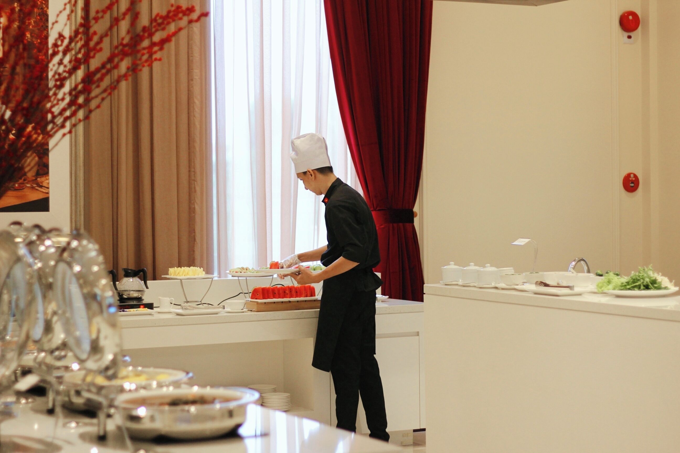 Una persona con divisa da chef sta preparando cibo in cucina, con una finestra, tende e un'area pranzo con piatti e un vaso.
