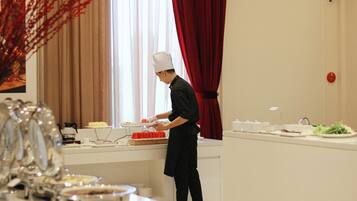 Una persona con divisa da chef sta preparando cibo in cucina, con una finestra, tende e un'area pranzo con piatti e un vaso.