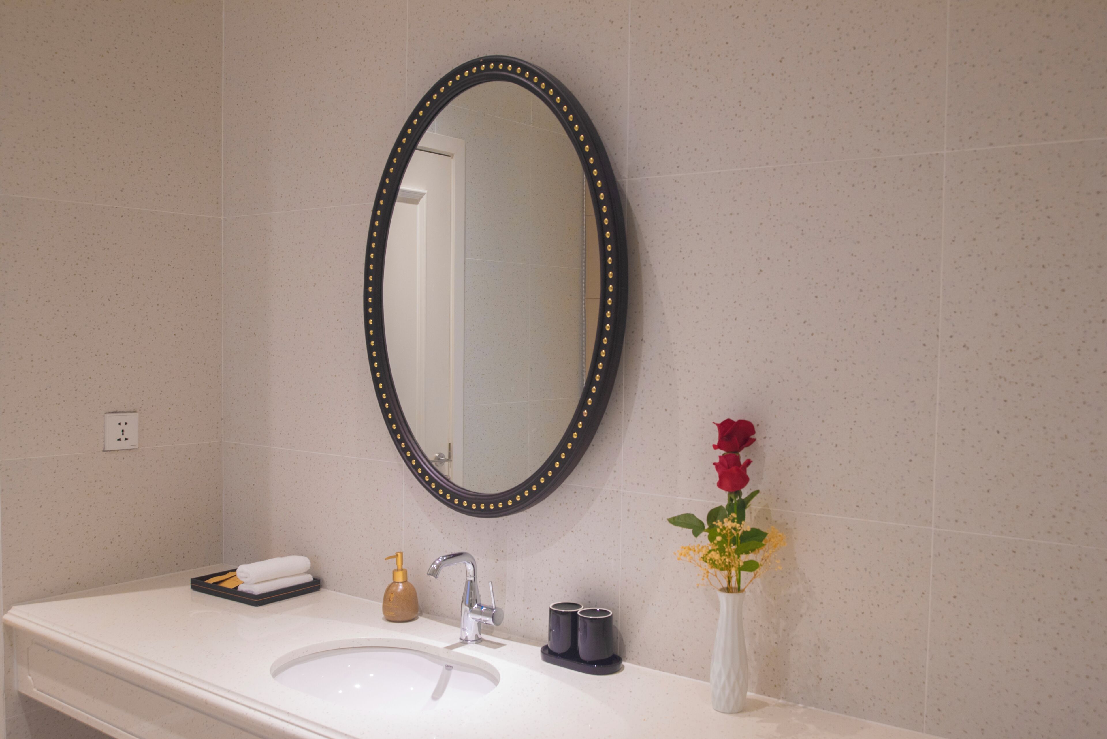 A bathroom with a white sink, a mirror with a black frame, a faucet, a soap dispenser, and a vase with flowers.