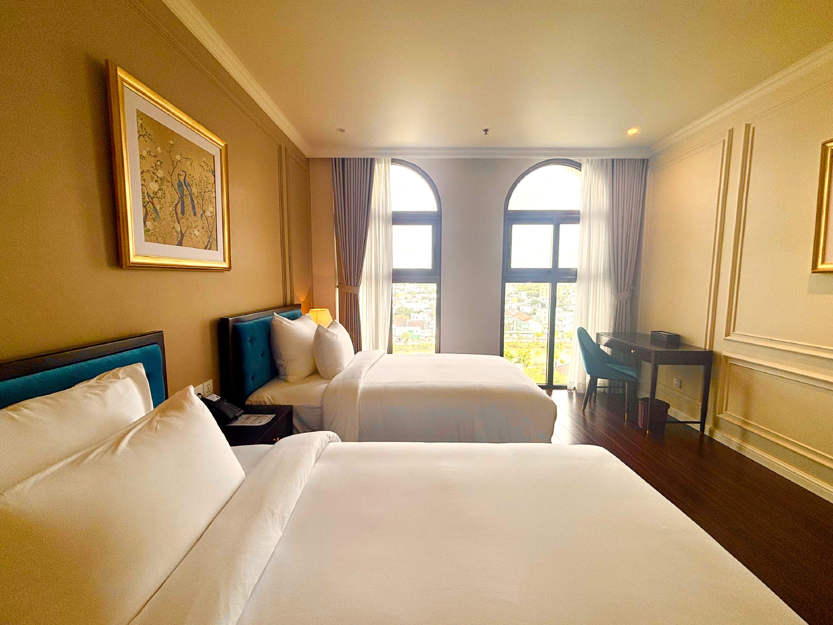 Habitación de hotel con dos camas, un escritorio y ventanas amplias.