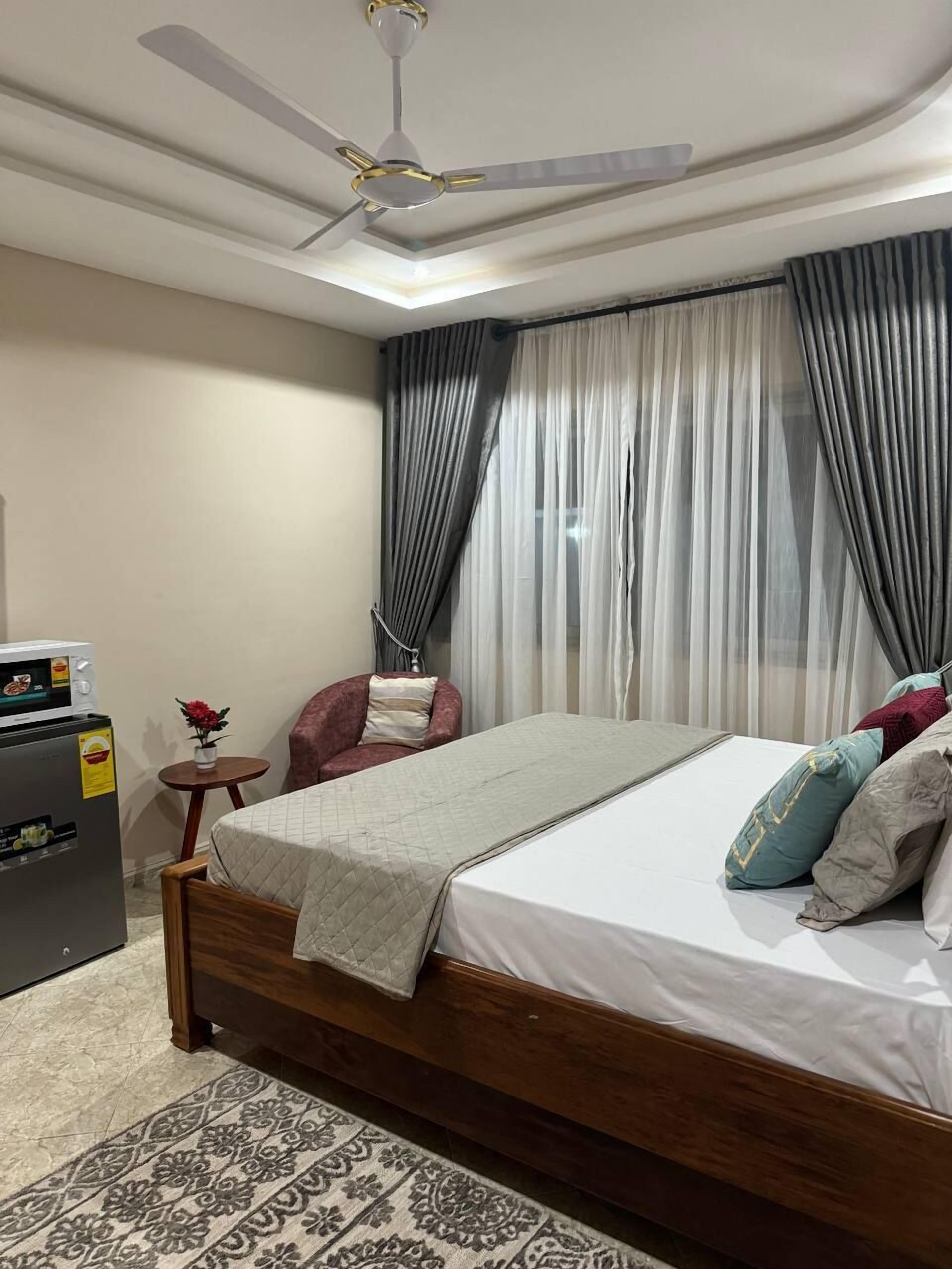 A bedroom with a bed, a ceiling fan, a small table with a vase, a chair, and a refrigerator.