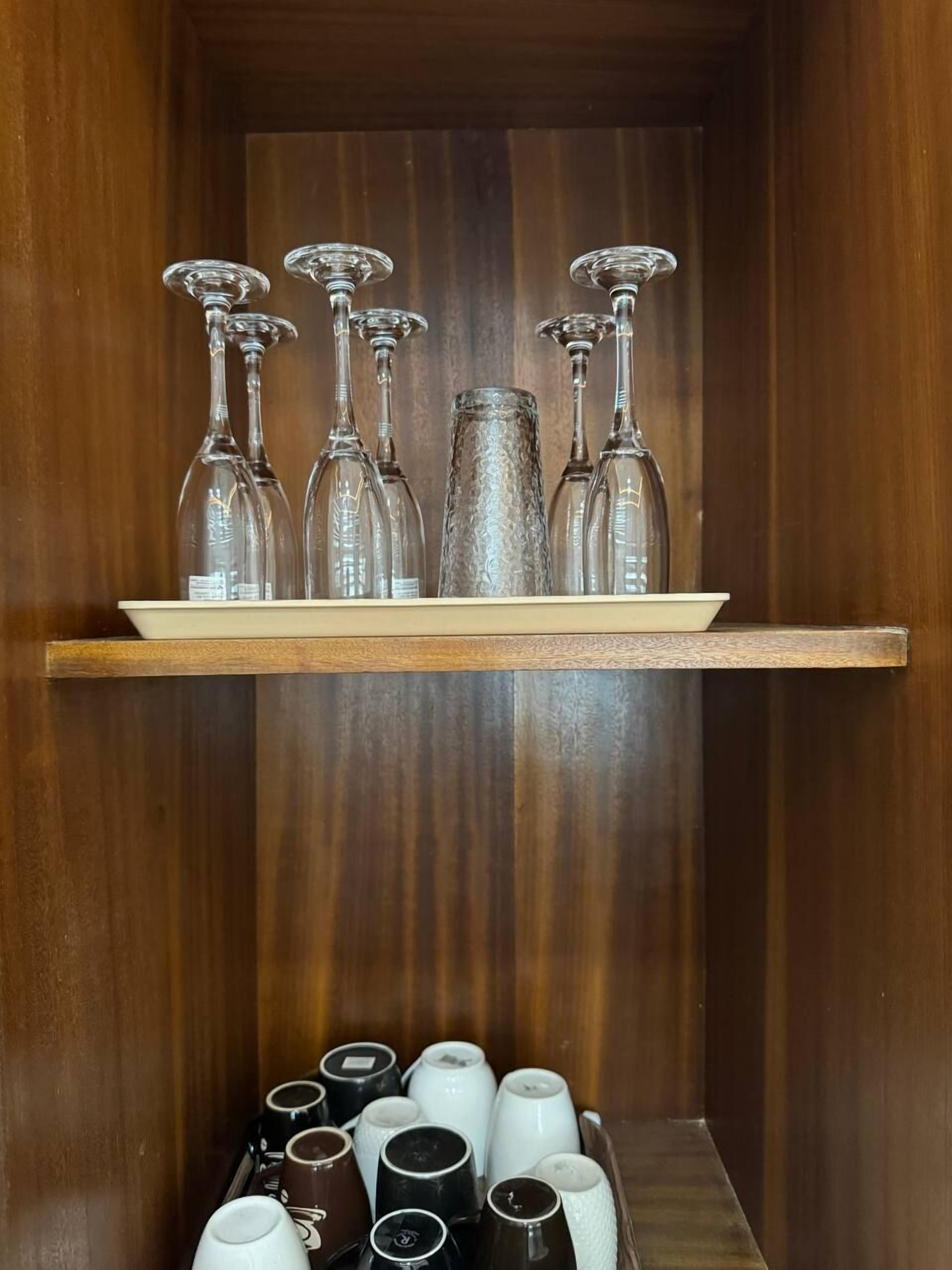 A wooden cabinet with a shelf holding several wine glasses and a tray below with various mugs.