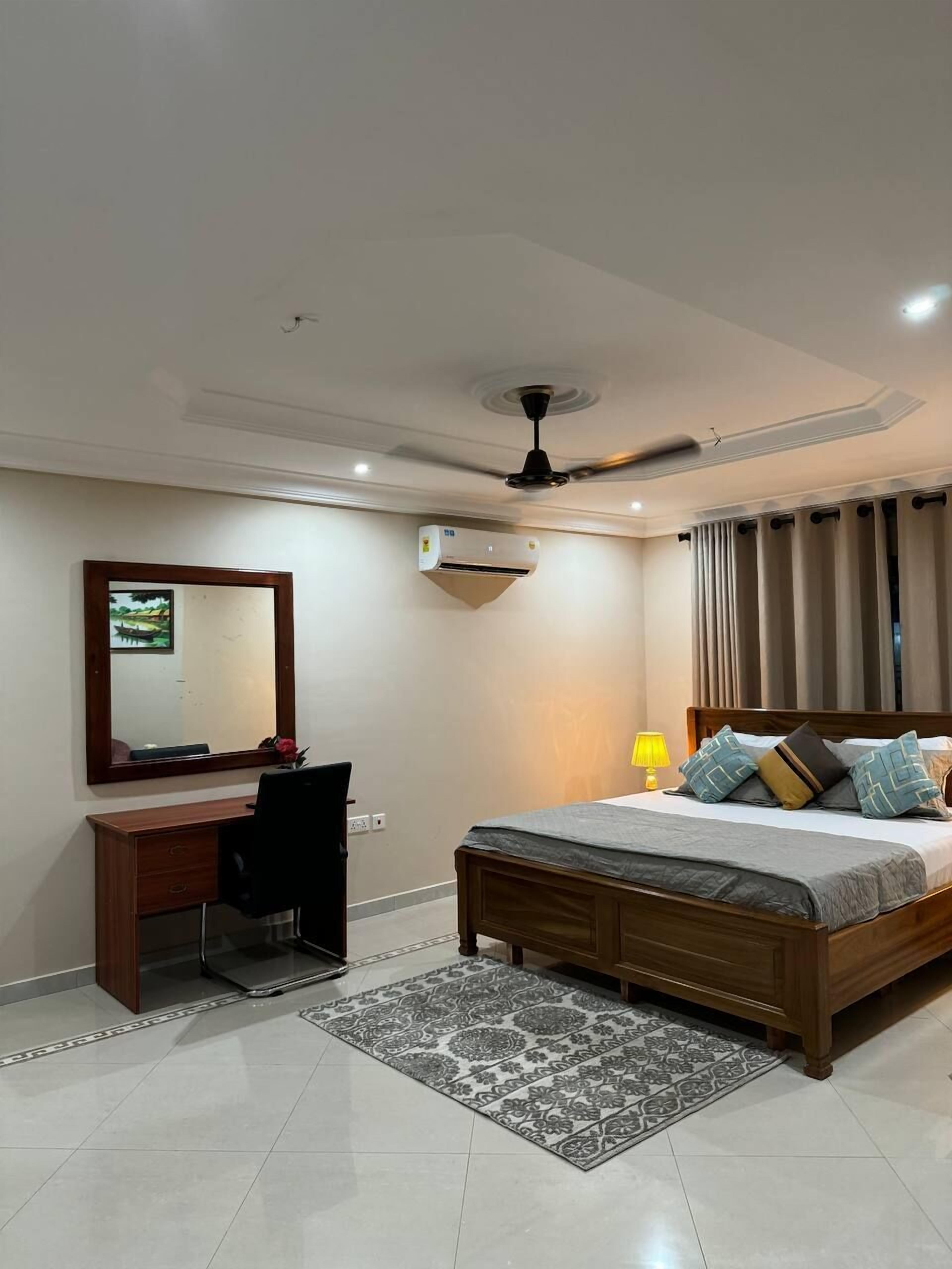 A hotel room with a bed, desk, chair, air conditioning unit, ceiling fan, and a mirror.