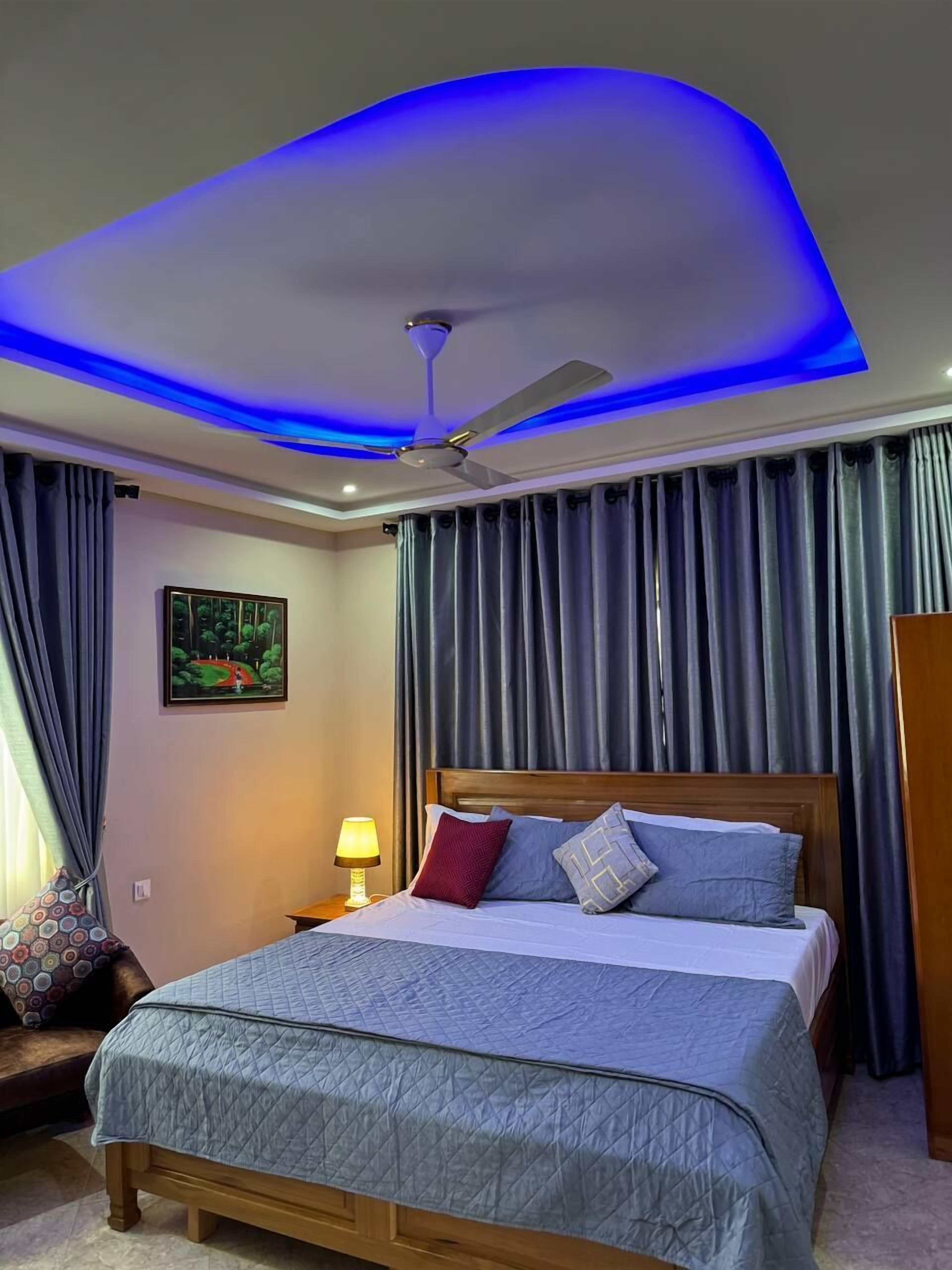 A hotel room with a bed, a ceiling fan, a nightstand, a lamp, and a painting on the wall.