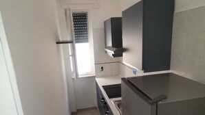 A narrow kitchen with a refrigerator, stove, and a window with blinds.