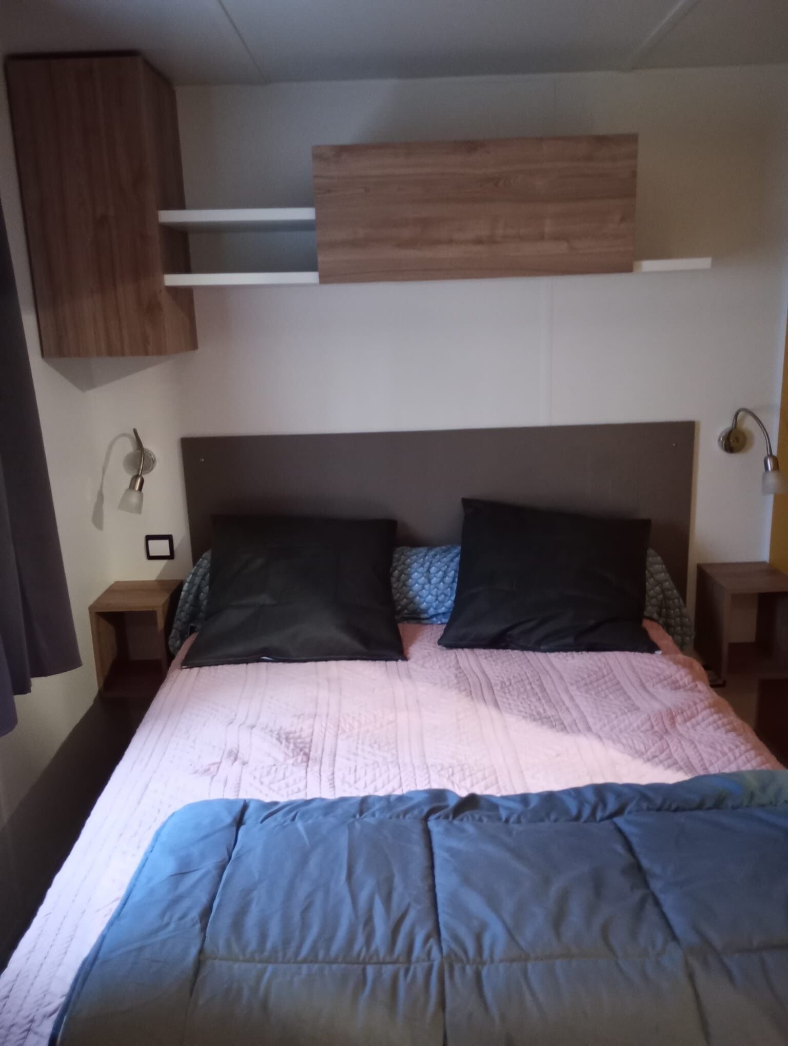 A bed with a headboard, two bedside tables, a wall-mounted shelf, and a wall-mounted lamp.