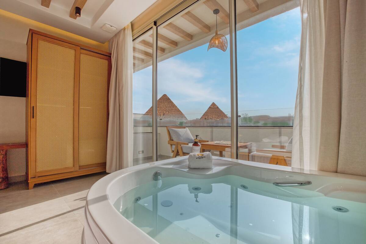 Une salle de bain moderne avec un jacuzzi entouré de parois en verre, des meubles en bois et une vue sur les pyramides.