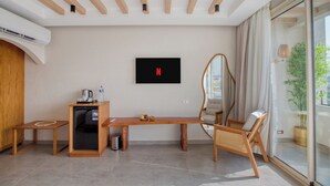 Une chambre moderne avec un plafond en bois, un téléviseur à écran plat fixé au mur, un petit coin cuisine, un banc en bois, une chaise en osier et une grande fenêtre donnant sur un paysage verdoyant.