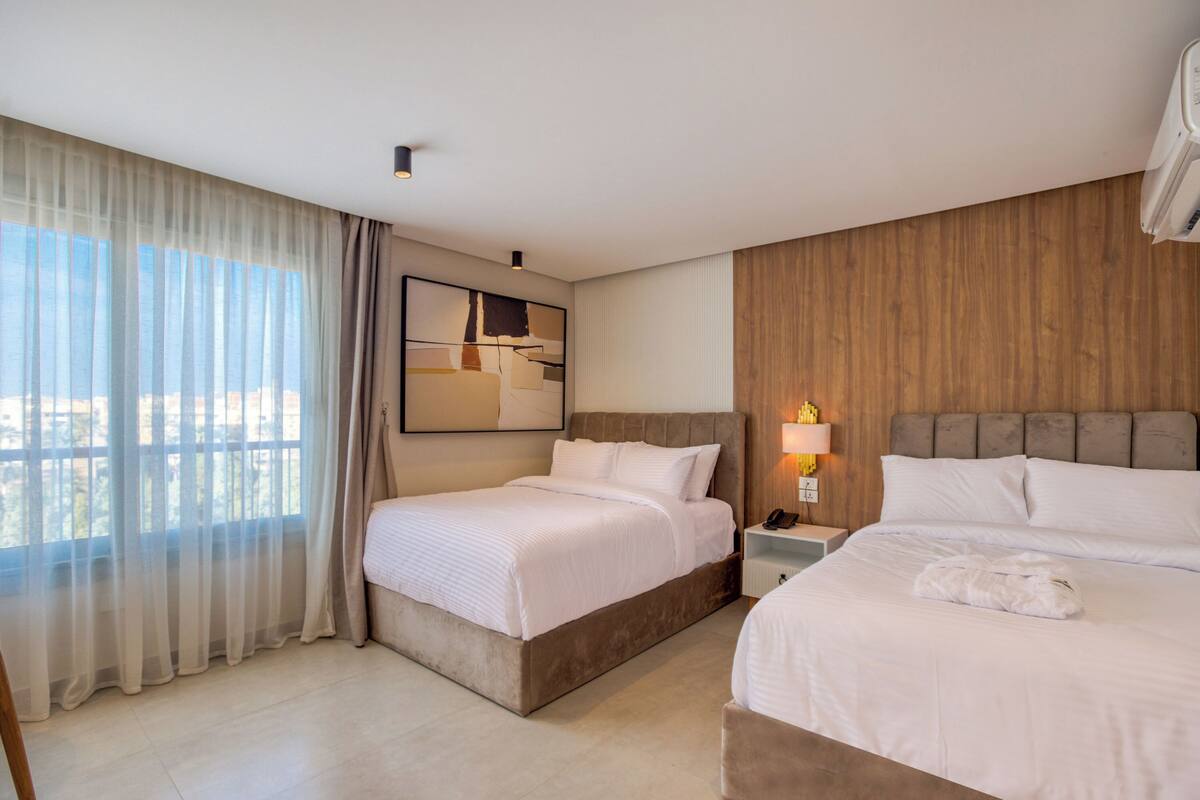 Une chambre d’hôtel avec deux lits, une grande fenêtre avec des rideaux légers, une tête de lit en bois, une table de chevet avec une lampe et une œuvre d’art accrochée au mur.