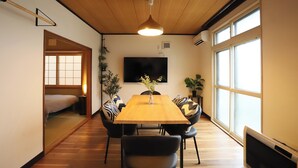 Una habitación con una mesa de comedor de madera, sillas negras, un televisor en la pared y ventanas amplias.