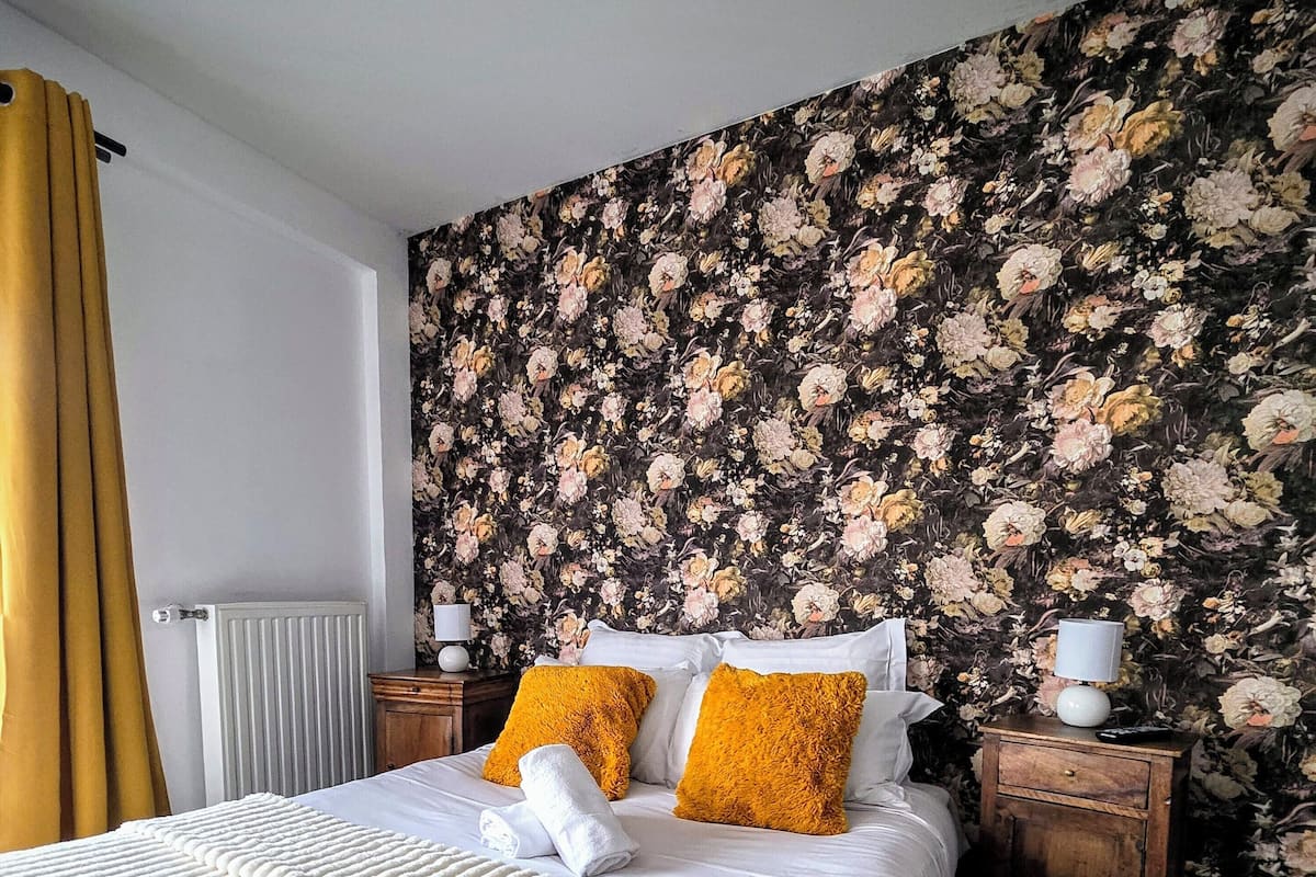 A hotel room with a floral wallpaper, a bed with white bedding and two orange pillows, a wooden nightstand with a lamp, and a radiator.