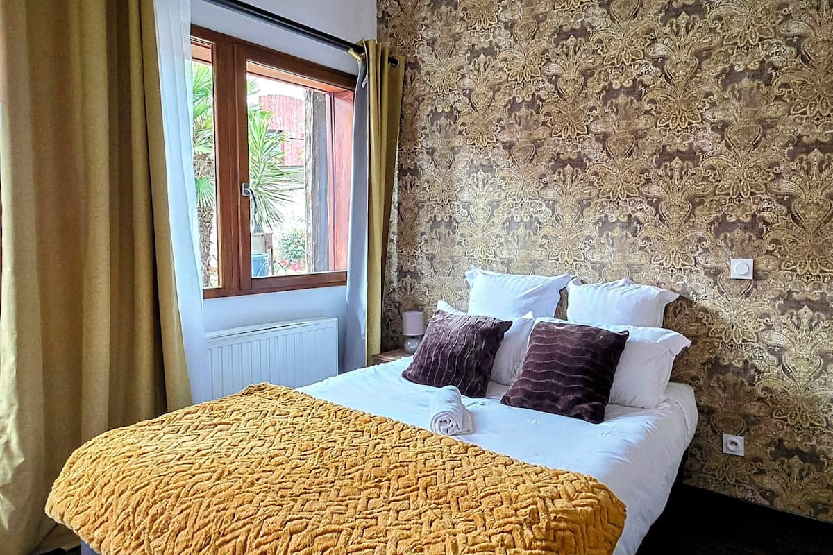 A bedroom with a bed, a window with curtains, patterned wallpaper, and a wooden ceiling beam.