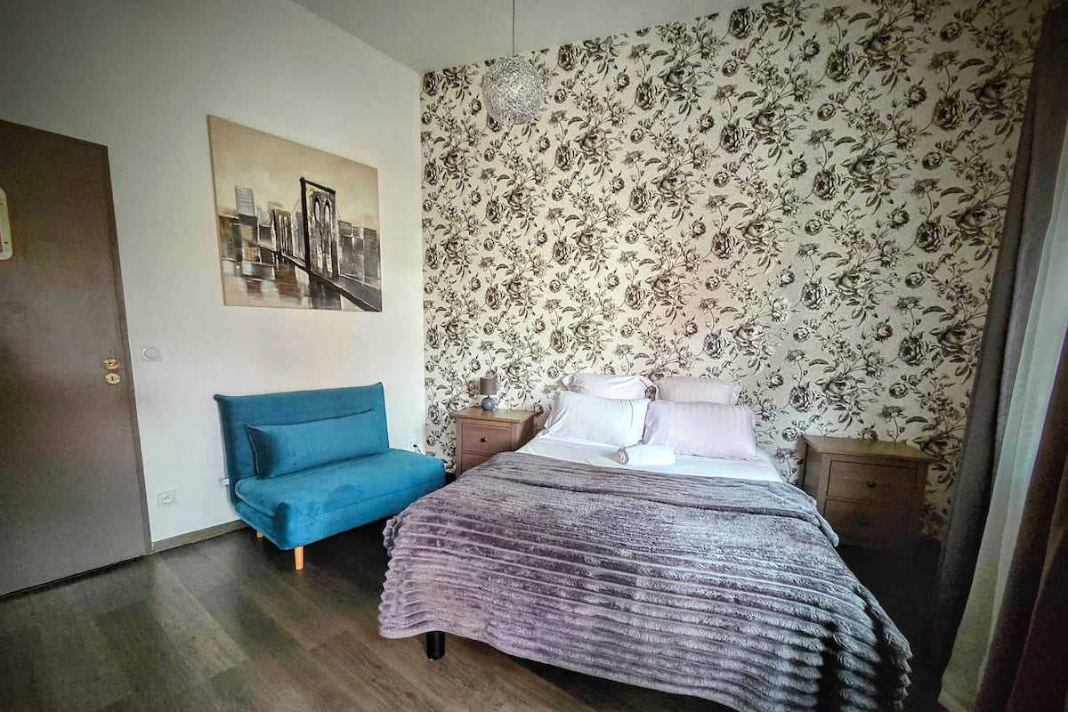 A bedroom with a bed, a blue sofa, a nightstand, a lamp, and a floral wallpaper.