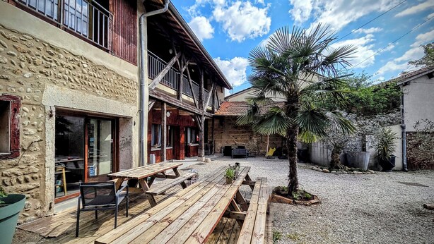 Un edificio rústico con fachada de piedra, balcones de madera y un patio de grava con una palmera.