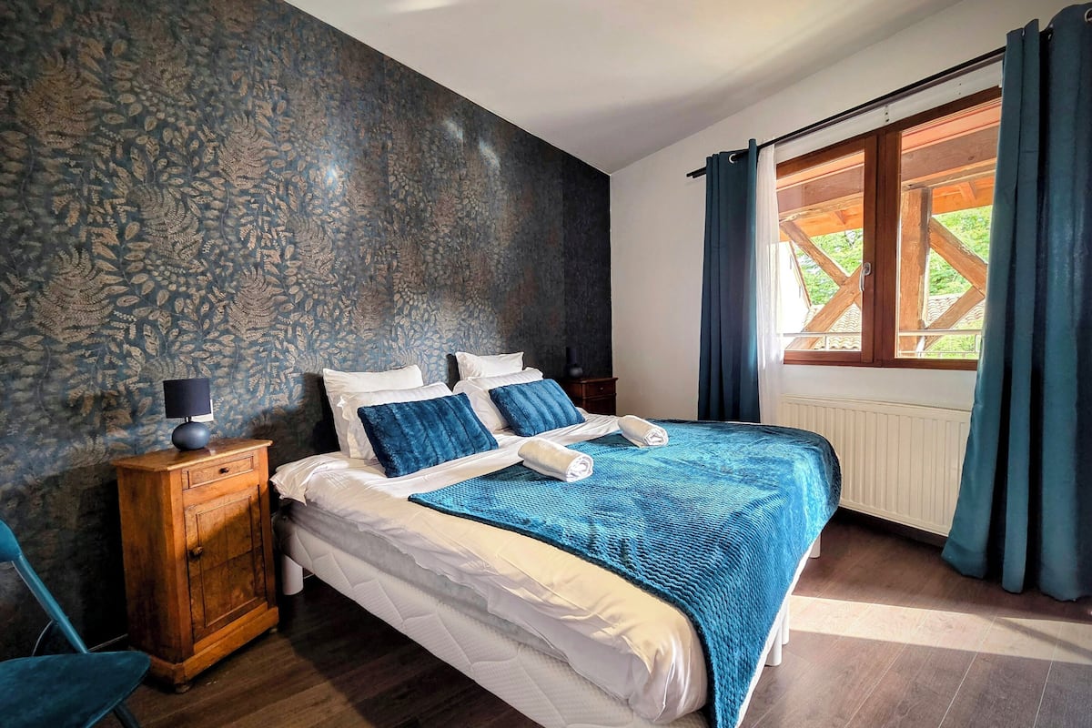 A bedroom with a bed, a nightstand, a lamp, a window with curtains, and a patterned wall.