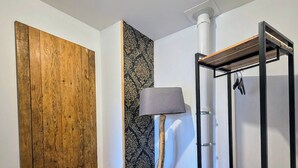 Una habitación con escritorio de madera, silla y lámpara con base de madera. Se observa una estantería con tres repisas y un perchero.
