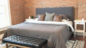 Un dormitorio con pared de ladrillo, cama con cabecera gris, una banca y alfombra estampada.